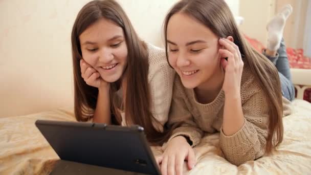 Retrato de dos adolescentes sonrientes que tienen videoconferencia en una tableta mientras están acostadas en la cama. — Vídeos de Stock