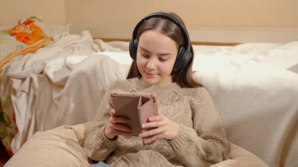 Smiling teenage girl listening music in headphones using tablet computer in her bedroom — Stock Video