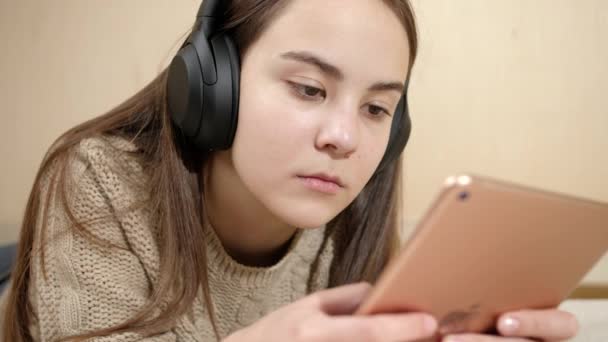 Brunetka nastolatka w słuchawki leżące na łóżku i za pomocą tabletu — Wideo stockowe