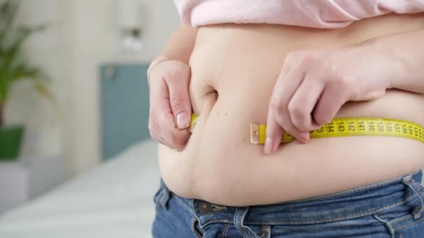 Νεαρή γυναίκα στέκεται στο υπνοδωμάτιο και μετράει τη μέση. Έννοια της δίαιτας, ανθυγιεινό τρόπο ζωής, υπέρβαρο και παχυσαρκία — Αρχείο Βίντεο
