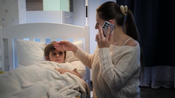 Молода мати торкається чола свого хворого сина, який лежить у ліжку, розмовляючи з лікарем по телефону. Концепція дитячої хвороби, хвороби та батьківського піклування — стокове відео