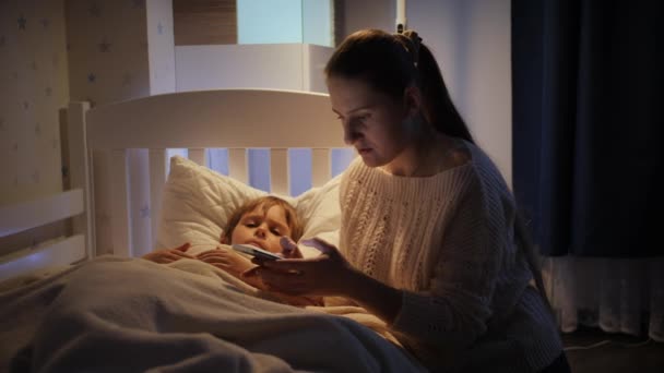 Νεαρή μητέρα ψάχνει για τον αριθμό τηλεφώνου του γιατρού στο smartphone για να τον καλέσει στο άρρωστο γιο της που βρίσκεται στο κρεβάτι. Έννοια της ασθένειας, της ασθένειας και της φροντίδας των γονέων — Αρχείο Βίντεο