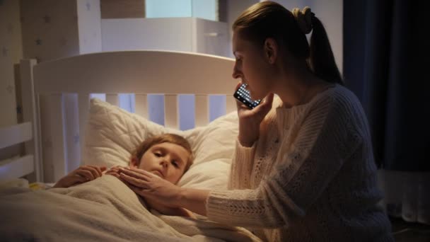 Malý nemocný chlapec leží v posteli vedle matky a telefonuje s doktorem. Koncepce dětské nemoci, nemoci a rodičovské péče — Stock video