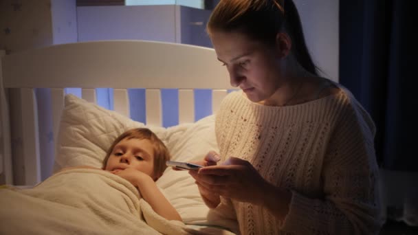 Joven madre preocupada escribiendo al médico en el teléfono inteligente junto a su pequeño hijo enfermo acostado en la cama. Concepto de enfermedad infantil, enfermedad y cuidado de los padres — Vídeo de stock