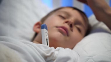 Annenin hasta oğluna nazikçe dokunup okşayarak yatakta yüksek ateşle yatması. Çocuk hastalığı, hastalık ve ebeveyn bakımı kavramı