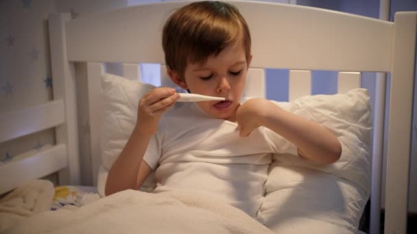 Malý nemocný chlapec leží v posteli a měří tělesnou teplotu pomocí teploměru. Pojem dětské nemoci, nemoci a zdravotní péče — Stock video