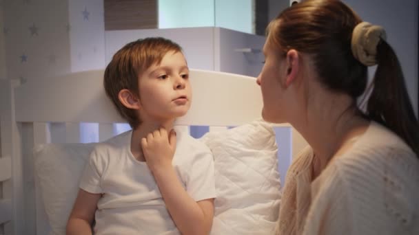 Caring mother checking and examining soring throat of her sick little son in bed. Concept of children illness, disease and parent care — Stock Video