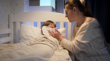 Hasta oğlunun yanında oturan ve burnunu kağıt mendile silen şefkatli genç bir anne. Çocuk hastalığı, hastalık ve ebeveyn bakımı kavramı