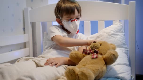 Leuke zieke jongen met beschermend medisch masker liggend in bed met teddybeer. Concept van het kind virus en kinderen bescherming tijdens coronavirus Covid-19 pandemie. — Stockvideo
