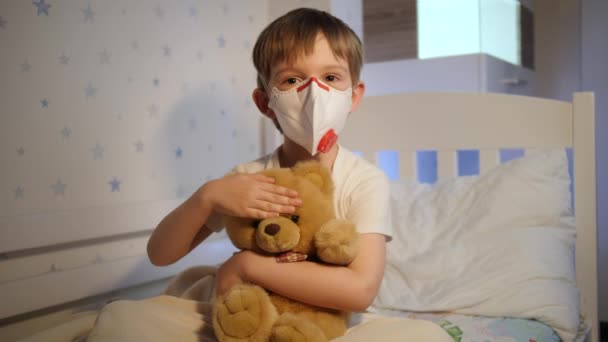 Netter kleiner Junge mit medizinischer Schutzmaske, der nachts seinen Teddybär im Bett hält und streichelt. Konzept des Kindervirus und Kinderschutzes während der Coronavirus-Covid-19-Pandemie. — Stockvideo