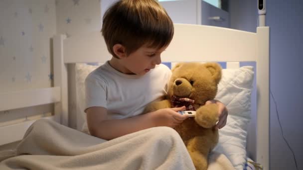 Leuke kleine jongen liggend in kraal en het meten van de temperatuur van zijn teddybeer speelgoed. Concept van het kind virus en kinderen bescherming tijdens coronavirus Covid-19 pandemie. — Stockvideo