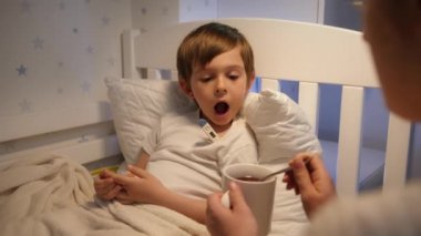 Anne hasta oğlunun yanına oturup ona ilaç ve sıcak çay veriyor. Çocuk hastalığı, hastalık ve ebeveyn bakımı kavramı