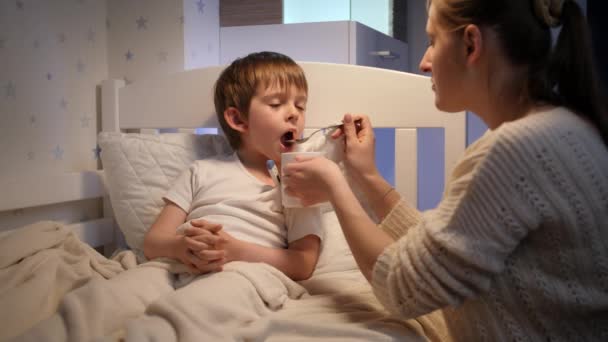 Η νεαρή μητέρα δίνει φάρμακα και σιρόπι από το κουτάλι στον άρρωστο γιο της ξαπλωμένο στο κρεβάτι. Έννοια της ασθένειας, της ασθένειας και της φροντίδας των γονέων — Αρχείο Βίντεο
