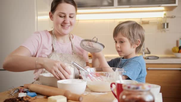 Kleine lachende jongen die zeef vasthoudt en moeder helpt met het maken van koekjesdeeg. Kinderen koken met ouders, kleine chef-kok, familie hebben tijd samen, huishoudelijke keuken. — Stockvideo