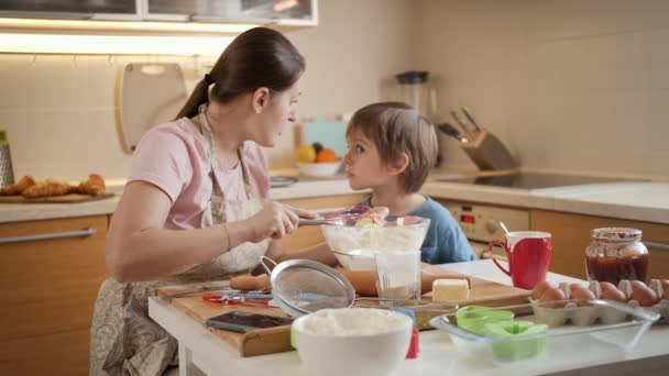 Ung kvinna pratar med sin lille son när hon bakar paj eller tårta hemma. Barn matlagning med föräldrar, liten kock, familj har tid tillsammans, kök. — Stockvideo