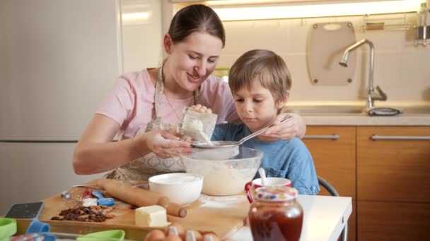 Eine junge lächelnde Mutter bringt ihrem Sohn bei, Mehl mit einem Sieb für die Teigzubereitung zu sieben. Kinder kochen mit Eltern, kleiner Koch, gemeinsame Zeit mit der Familie, häusliche Küche. — Stockvideo