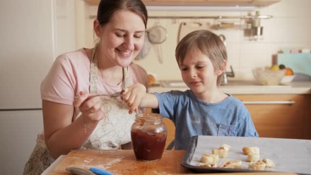 Χαρούμενη χαμογελαστή μητέρα με τον μικρό γιο να τρώει γλυκιά μαρμελάδα από το βάζο ενώ ψήνει μπισκότα στο σπίτι. Παιδιά μαγείρεμα με τους γονείς, λίγο σεφ, οικογένεια έχοντας χρόνο μαζί, οικιακή κουζίνα. — Αρχείο Βίντεο