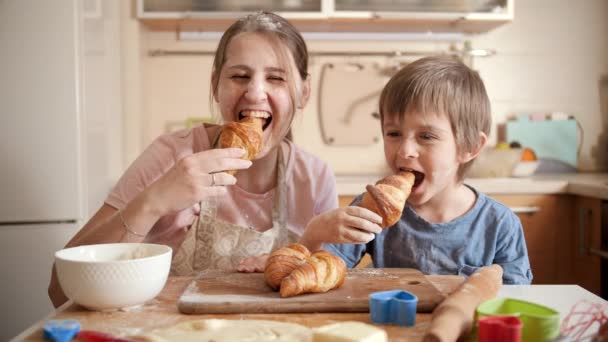 Glad leende pojke med mor täckt av mjöl äta välsmakande croissant efter bakning hemma. Barn matlagning med föräldrar, liten kock, familj har tid tillsammans, kök. — Stockvideo