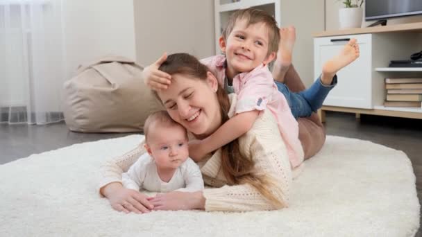 Leuke jongen die zijn moeder en broertje knuffelt liggend op tapijt in de woonkamer. Ouderschap, kindergeluk en familierelatie — Stockvideo