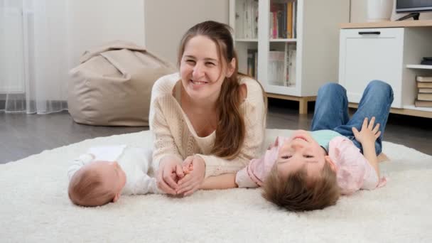 Młoda uśmiechnięta matka leżąca ze swoim małym dzieckiem i starszymi synami na dywanie w salonie. Rodzicielstwo, szczęście dzieci i relacje rodzinne — Wideo stockowe
