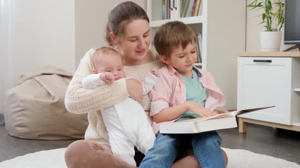 Jovem mãe amorosa sentada com seus filhos na sala de estar e lendo livro de histórias. Paternidade, felicidade das crianças e relacionamento familiar — Vídeo de Stock