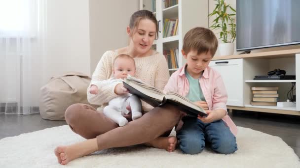 Gülümseyen anne yerde oturuyor ve büyük ve küçük oğullarına masal kitabı okuyor. Ebeveynlik, çocuklar mutluluk ve aile ilişkisi — Stok video