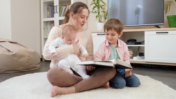 Счастливая семья с детьми, сидящими на полу в гостиной и читающими книжку. Родители, дети счастье и семейные отношения — стоковое видео