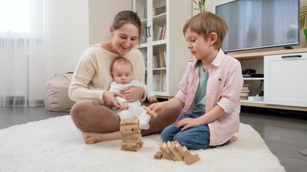 Fiatal anya fogja a kisfiút és játszik az idősebb fiával fablokkokban. Szülői, gyermeki boldogság és családi kapcsolat — Stock videók