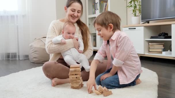 Счастливые улыбающиеся мальчики и мама играют с деревянными игрушечными блоками на полу в гостиной. Родители, дети счастье и семейные отношения — стоковое видео