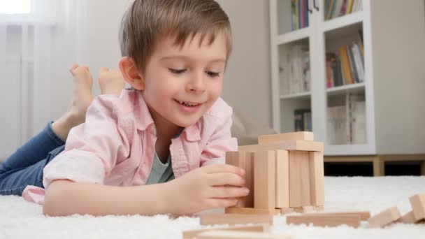 Ragazzo sorridente sdraiato sul pavimento e edificio casa o torre da blocchi di legno. Concetto di educazione dei bambini e giochi a casa — Video Stock