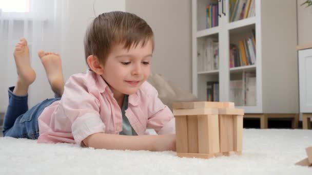 Leuke lachende jongen liggend op de vloer en kijkend naar speelgoed huis dat hij bouwde van speelgoed houten blokken. Concept van kinderopvoeding en spelletjes thuis — Stockvideo