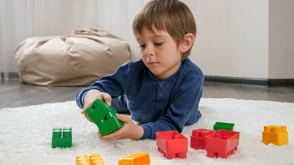 Милый мальчик лежит на мягком ковре и играет с красочными игрушечными блоками. Концепция детского образования и игр на дому — стоковое фото