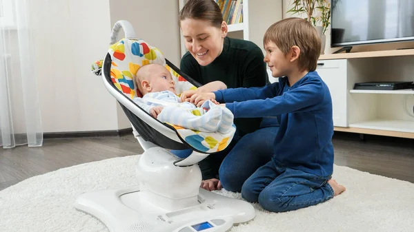 Ağabeyi ve gülümseyen annesi elektrikli sandalyede sallanan küçük bebekle oynuyorlar. Çocuk gelişimi ve mutlu çocukluk — Stok fotoğraf