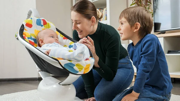 Lindo niño sonriente meciéndose en la silla y mirando a la madre y al hermano mayor. Desarrollo infantil y infancia feliz — Foto de Stock