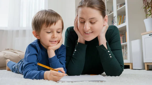 Jonge moeder kijkt naar haar zoontje die huiswerk maakt op tapijt in de woonkamer. Concept van kinderopvoeding, ontwikkeling en gelukkig ouderschap. — Stockfoto