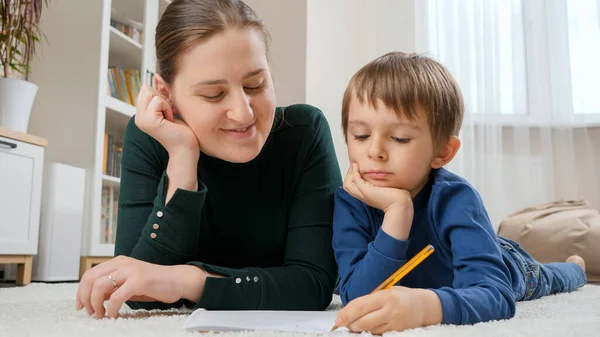 小さな男の子と若い母親がリビングルームに床に横たわっており、コピーブックで宿題をしています。子供の教育、開発と幸せな子育ての概念. — ストック写真