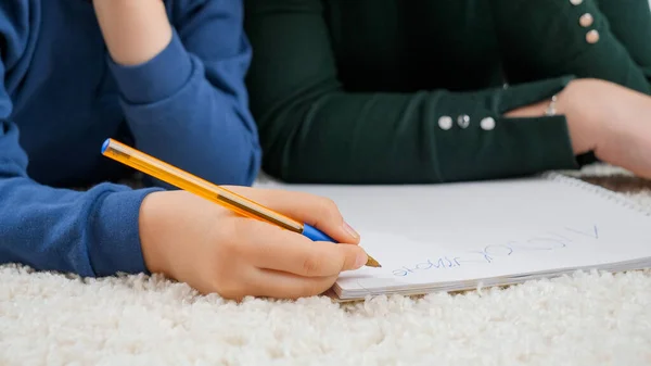 Primer plano de niño pequeño escribiendo con pluma en copybook mientras está acostado en la alfombra en la sala de estar. Concepto de educación infantil, desarrollo —  Fotos de Stock