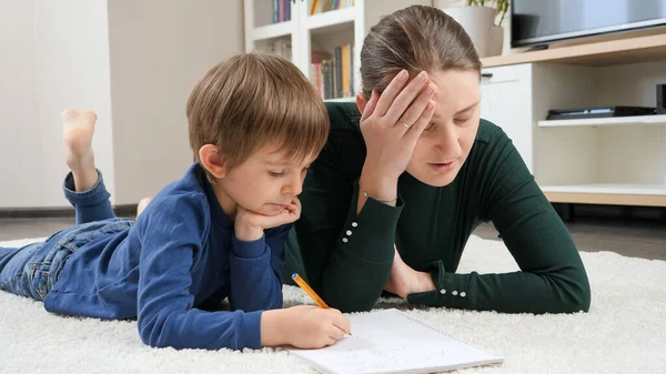 Dispelased angry mother lying next to little son doing homework on carpet — Stock Photo, Image