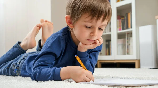 Porträtt av en söt liten pojke som gör läxor på matta i vardagsrummet. Begreppet hemundervisning och utveckling av barn. — Stockfoto