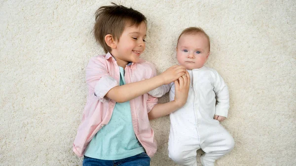 Felice ragazzo sorridente sdraiato con il suo fratellino sul tappeto e divertirsi. Felicità dei bambini e relazioni familiari. — Foto Stock