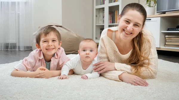 Szczęśliwe dzieci z matką leżącą na dywanie i uśmiechniętą przed kamerą. — Zdjęcie stockowe