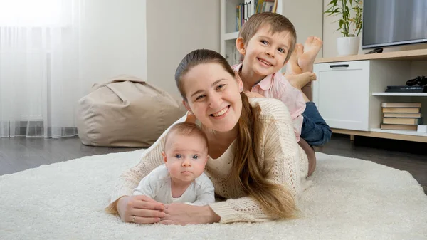 Bambini sorridenti felici che si abbracciano e si divertono con la madre sul tappeto in soggiorno. Genitorialità, felicità dei bambini e relazione familiare — Foto Stock