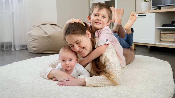 Милый мальчик обнимает мать и младшего брата, лежащих на ковре в гостиной. Родители, дети счастье и семейные отношения — стоковое фото