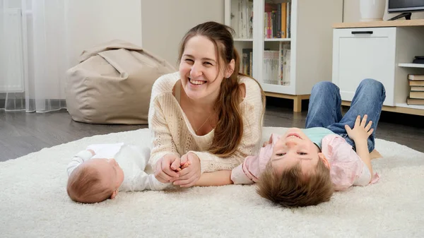 Ung leende mamma ligger med sin lilla bebis och äldre söner på matta i vardagsrummet. Föräldraskap, barns lycka och familjeförhållande — Stockfoto