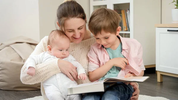 Usmívající se žena objímá své dítě a staršího syna, zatímco čte knihu příběhů v obýváku. Rodičovství, dětské štěstí a rodinný vztah — Stock fotografie