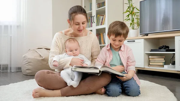 Uśmiechnięta matka, siedząca na podłodze i czytająca opowiadania swoim starszym i młodszym synom. Rodzicielstwo, szczęście dzieci i relacje rodzinne — Zdjęcie stockowe