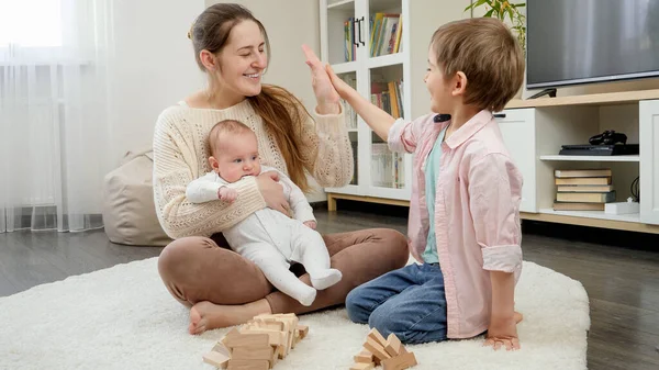 Счастливые улыбающиеся мальчики и мама играют с деревянными игрушечными блоками на полу в гостиной. Родители, дети счастье и семейные отношения — стоковое фото