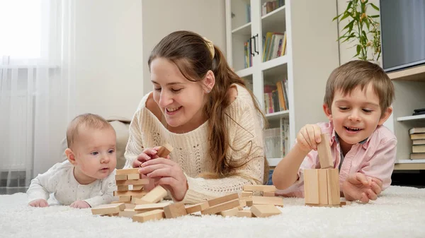 Маленький ребенок, мама и старший брат играют с деревянными блоками на полу в гостиной. Родители, дети счастье и семейные отношения — стоковое фото
