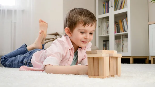 Roztomilý usměvavý chlapec ležící na podlaze a dívající se na domeček z hraček, který postavil z dřevěných bloků. Koncepce dětské výchovy a hry doma — Stock fotografie