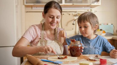 Gülümseyen anne ve küçük oğlu pasta ya da tatlı reçel ile pasta dolduruyor. Çocuklar aileleriyle yemek pişiriyor, küçük aşçı, aile birlikte vakit geçiriyor, ev mutfağı yapıyor.